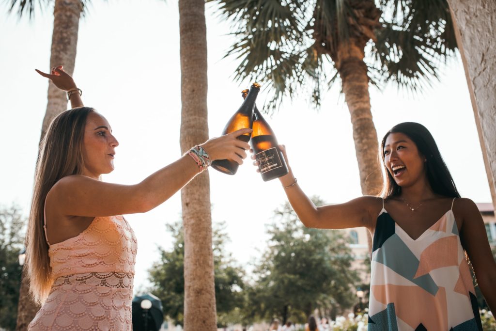 English woman and asian woman toasting wine bottles with palm trees.